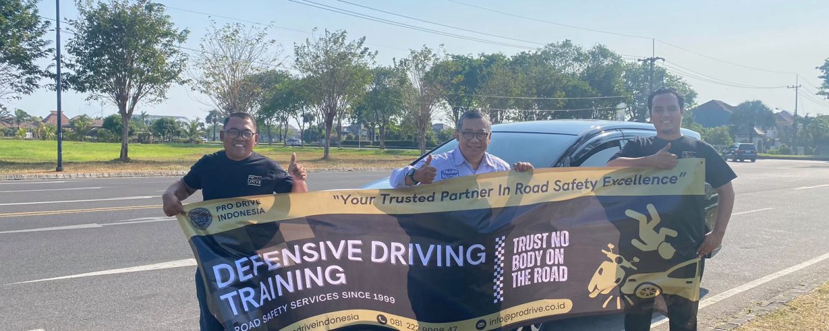 Tiga orang berfoto sambil memegang banner Defensive Driving Training Pro Drive Indonesia di tepi jalan.