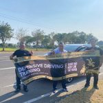 Tiga orang berfoto sambil memegang banner Defensive Driving Training Pro Drive Indonesia di tepi jalan.